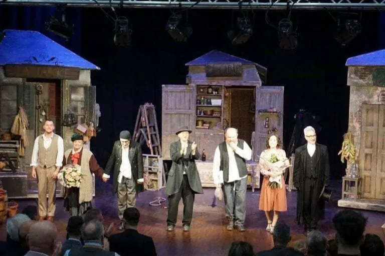 Oamenii stau pe o scenă de teatru în fața unui public, purtând diverse costume, lângă decoruri care reprezintă structuri de lemn.