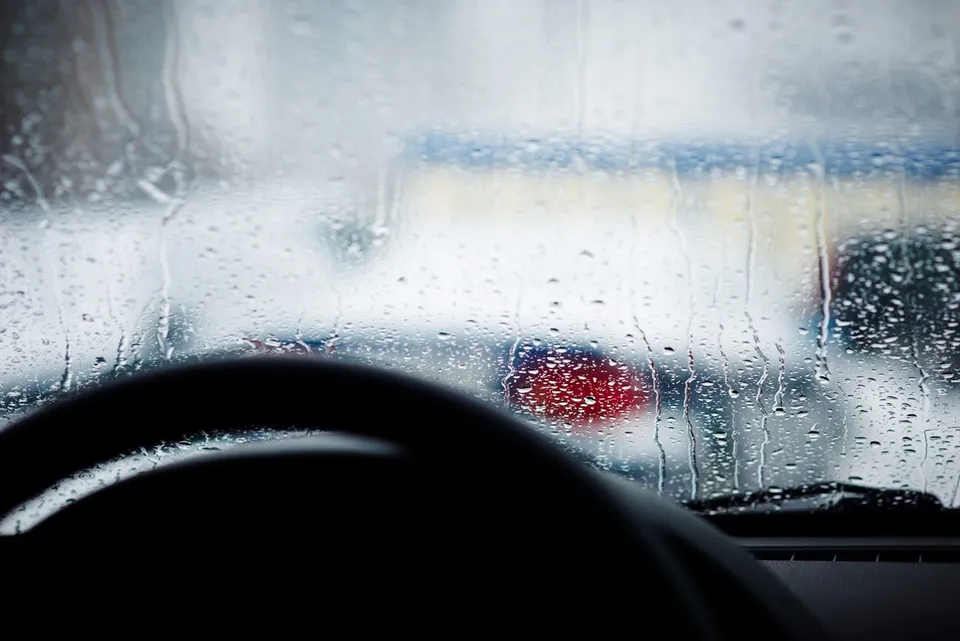 Picături de apă și condens acoperă un parbriz de autovehicul, prin care se observă silueta difuză a unei mașini albe cu lumini roșii.