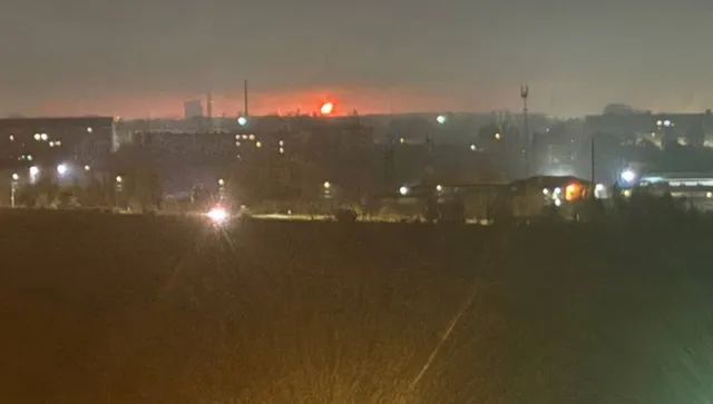 O lumină portocalie strălucitoare este vizibilă pe linia orizontului deasupra unor siluete de clădiri și structuri industriale sub un cer nocturn.