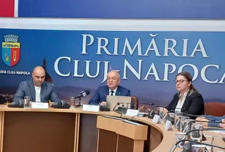 Trei persoane stau la o masă curbată în fața unui panou albastru pe care scrie Primăria Cluj-Napoca.