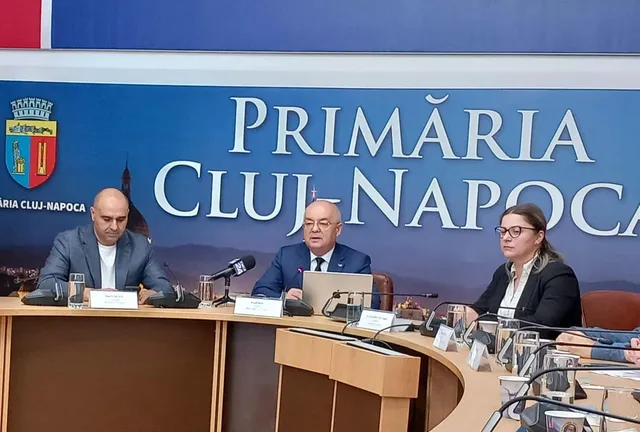 Trei persoane stau la o masă curbată în fața unui panou albastru pe care scrie Primăria Cluj-Napoca.