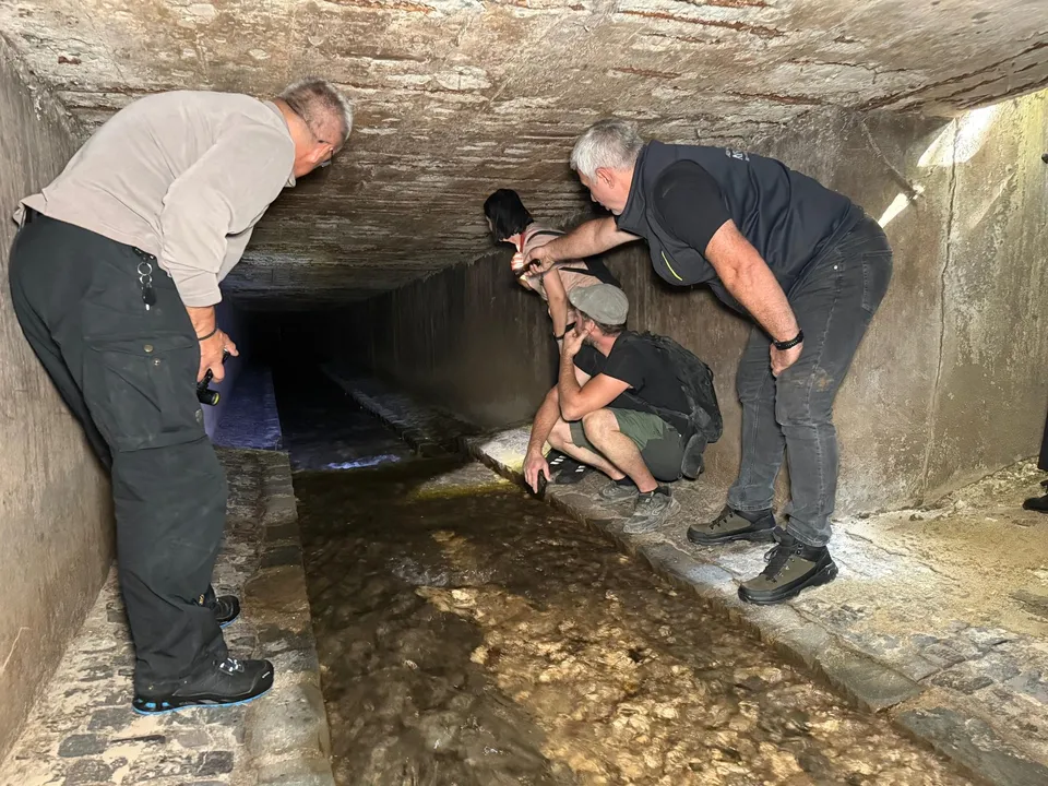 Cinci bărbați privesc într-un canal îngust de piatră sub o structură de beton cu lumină difuză.