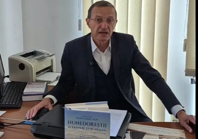 Un bărbat cu ochelari, purtând sacou și vestă, stă la o masă lângă un imprimant și o carte intitulată Hunedoreștii o familie europeană.