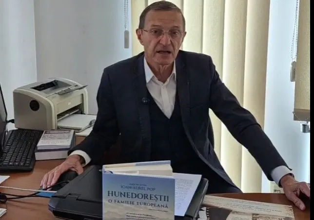 Un bărbat cu ochelari, purtând sacou și vestă, stă la o masă lângă un imprimant și o carte intitulată Hunedoreștii o familie europeană.