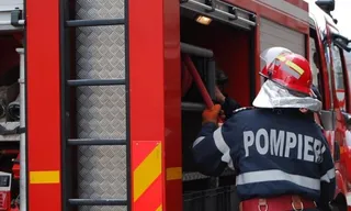 Un pompier cu costum de protecție și cască roșie, cu inscripția POMPIERI pe spate, este filmat din spate în timp ce accesează echipamentul unui camion de stingere a incendiilor roșu.