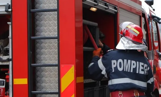 Un pompier cu costum de protecție și cască roșie, cu inscripția POMPIERI pe spate, este filmat din spate în timp ce accesează echipamentul unui camion de stingere a incendiilor roșu.