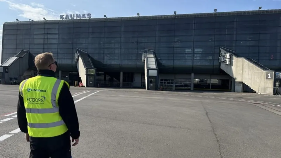 Un bărbat cu vestă reflectorizantă galbenă și neagră privește spre o clădire mare de terminal cu fațadă de sticlă și inscripția Kaunas.