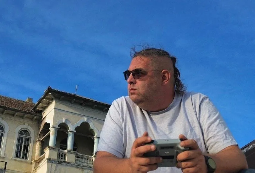 Un bărbat cu o coafură tip mohawk și ochelari de soare poartă un tricou alb și ține un obiect gri în mâini în fața unei clădiri.