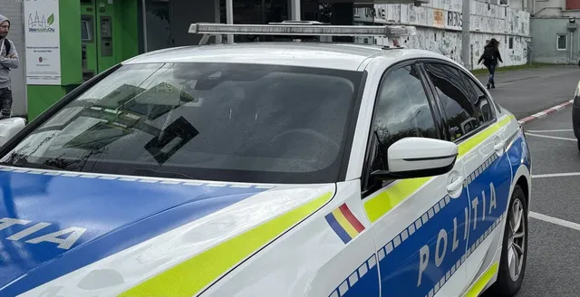 Un autoturism de poliție alb cu dungi albastre și galbene este parcat pe o stradă lângă un trotuar.