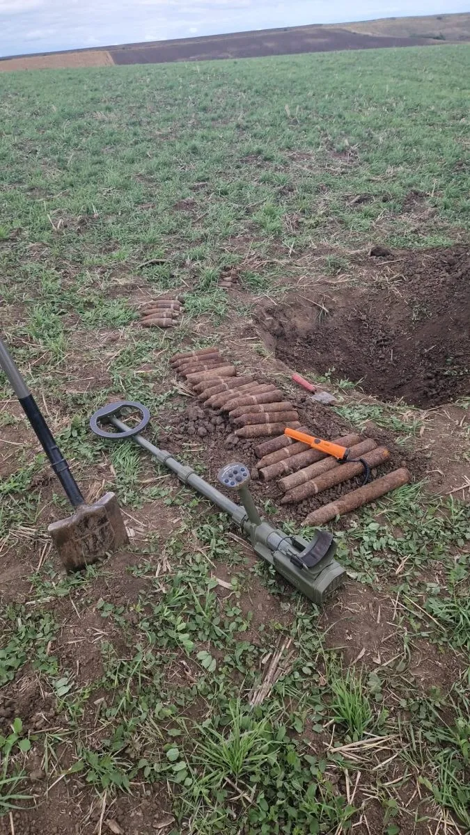 Un detector de metale și o lopată sunt așezate pe iarbă lângă mai multe obiecte cilindrice metalice dispuse pe sol lângă un șanț de pământ.