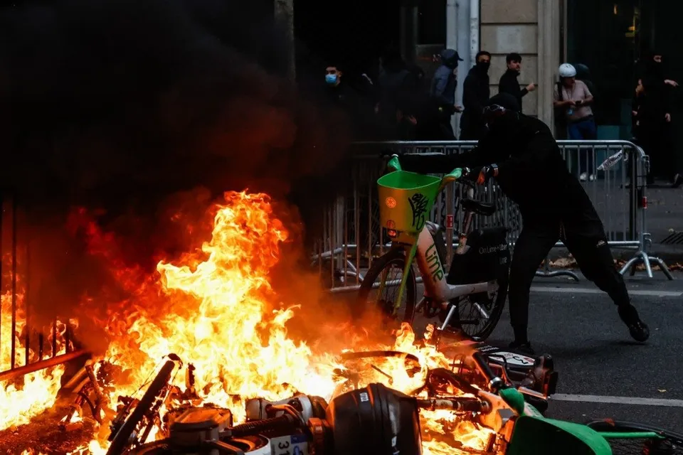 O persoană îmbrăcată în negru stă lângă un bicicletă verde și un foc intens care arde pe carosabil.