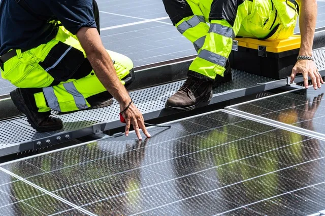 Două persoane purtând pantaloni fluorescenți de lucru se apleacă peste un rând de panouri fotovoltaice instalate pe o suprafață plată.