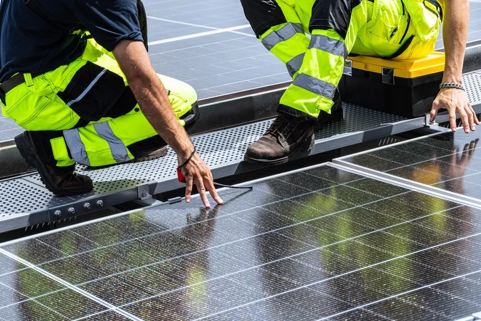 Două persoane purtând pantaloni fluorescenți de lucru se apleacă peste un rând de panouri fotovoltaice instalate pe o suprafață plată.