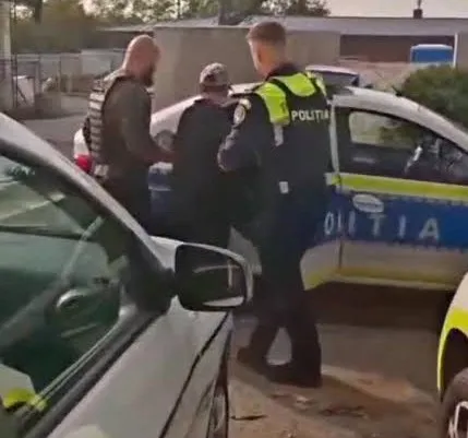 Un polițist în uniformă neagră și galbenă stă lângă o mașină albastră cu inscripția POLIȚIA, în timp ce alți doi bărbați sunt prezenți lângă vehicul.