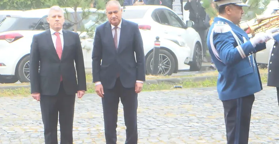 Trei bărbați stau pe o stradă pavată, doi purtând costume și unul purtând o uniformă albastră cu șapcă.