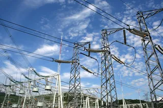 Mai multe turnuri metalice de înaltă tensiune susțin cabluri electrice sub un cer albastru cu nori albi.