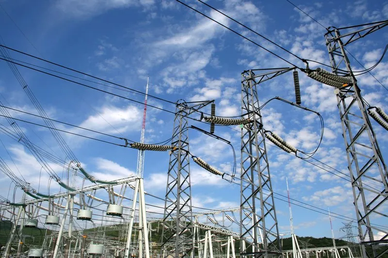 Mai multe turnuri metalice de înaltă tensiune susțin cabluri electrice sub un cer albastru cu nori albi.
