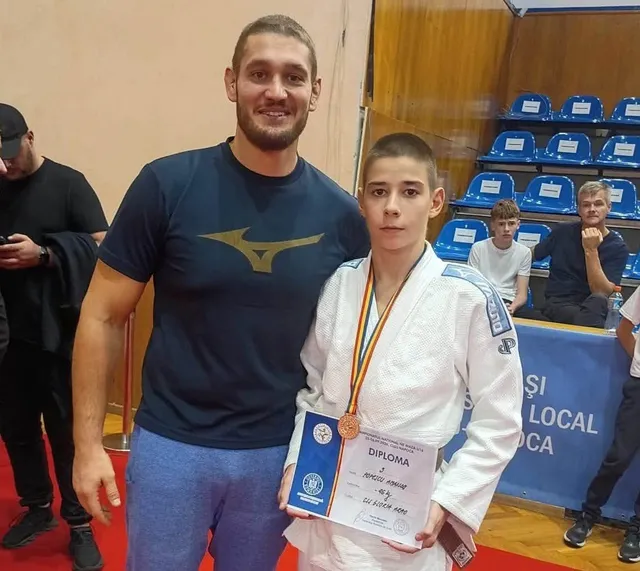 Un băiat într-un costum alb de judo poartă o medalie și ține o diplomă, stând lângă un bărbat cu tricou închis la culoare.