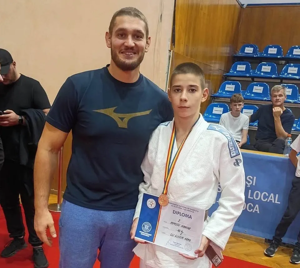 Un băiat într-un costum alb de judo poartă o medalie și ține o diplomă, stând lângă un bărbat cu tricou închis la culoare.