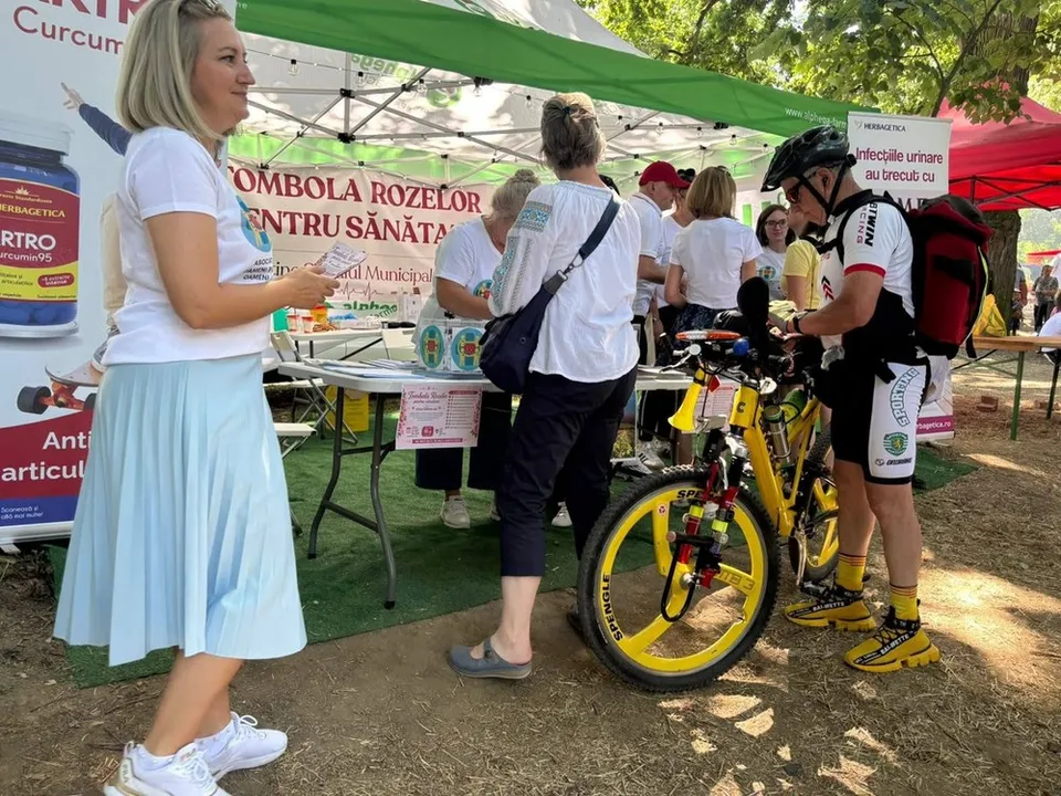 O femeie cu fustă albastră și tricou alb ține un bilet lângă un stand de eveniment, în timp ce un biciclist stă lângă bicicleta sa galbenă.