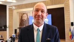 Un bărbat cu părul rare, purtând sacou închis la culoare, cămașă albă și cravată turcoaz, stă în fața unui fundal cu un tablou și un ecran albastru.
