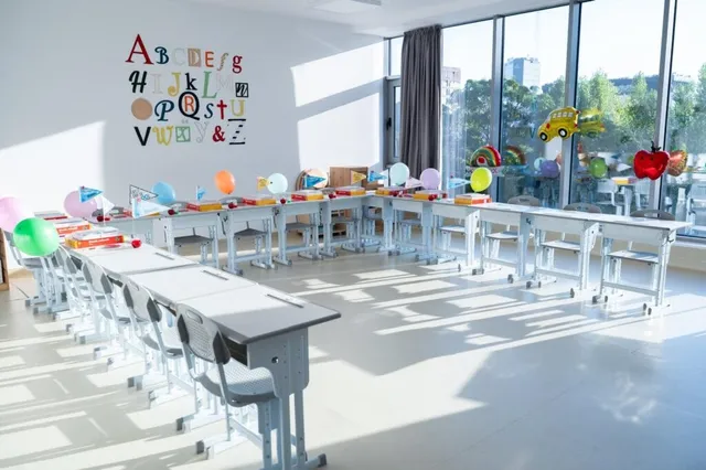 O sală de clasă luminoasă conține rânduri de bănci gri și scaune albe, fiind decorată cu baloane colorate și litere ale alfabetului pe perete.