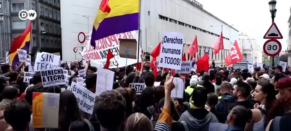 O mulțime de oameni ridică diverse pancarte și steaguri colorate în fața unei clădiri albe sub un cer deschis.