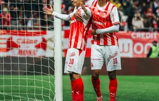 Doi jucători de fotbal în echipamente alb-roșii sărbătoresc pe un teren de fotbal lângă o plasă de porți.