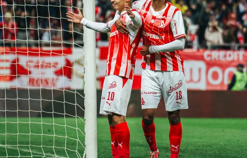 Doi jucători de fotbal în echipamente alb-roșii sărbătoresc pe un teren de fotbal lângă o plasă de porți.