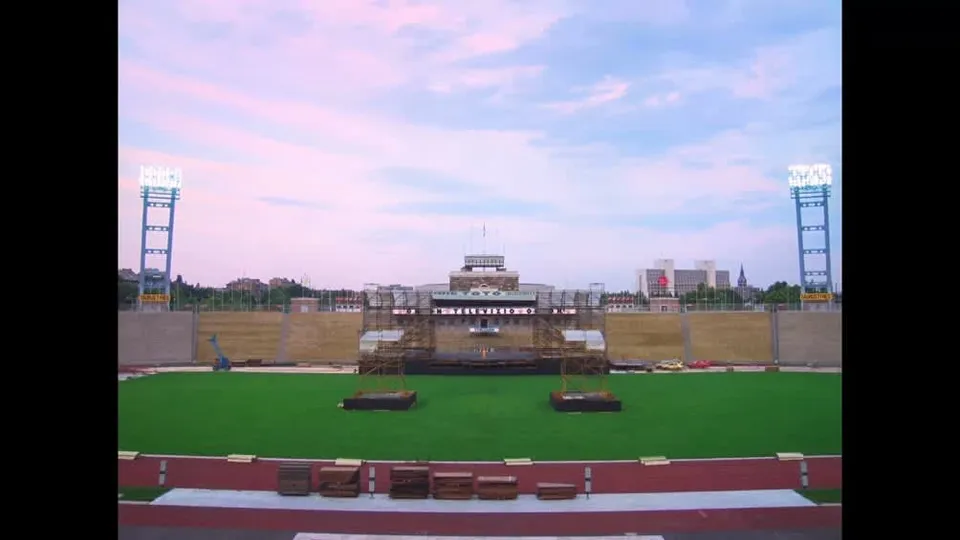 O scenă de concert este montată pe un teren verde, flancată de două turnuri de iluminat înalte sub un cer cu nuanțe roz și albastru.