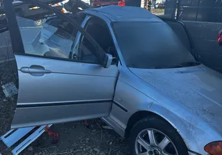 Un autoturism argintiu cu portiera șoferului deschisă este parcat pe un teren cu pietriș lângă un zid gri.