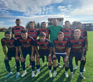 Un grup de fotbaliști în echipamente de sport roșii și negre pozează pe un teren de fotbal sub un cer cu nori.