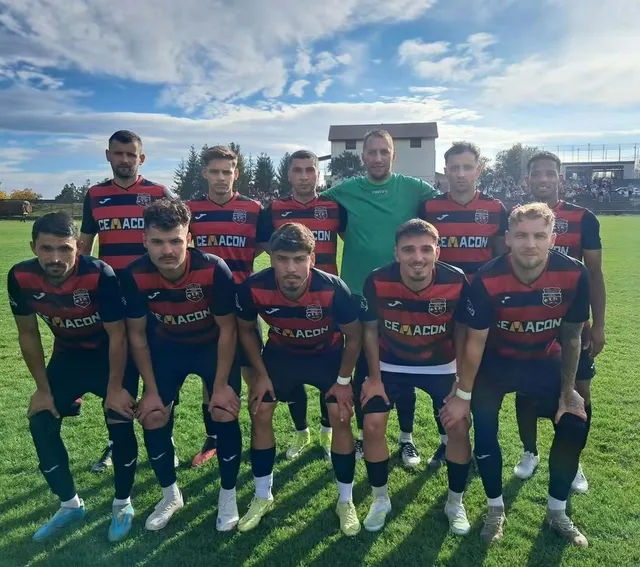 Un grup de fotbaliști în echipamente de sport roșii și negre pozează pe un teren de fotbal sub un cer cu nori.