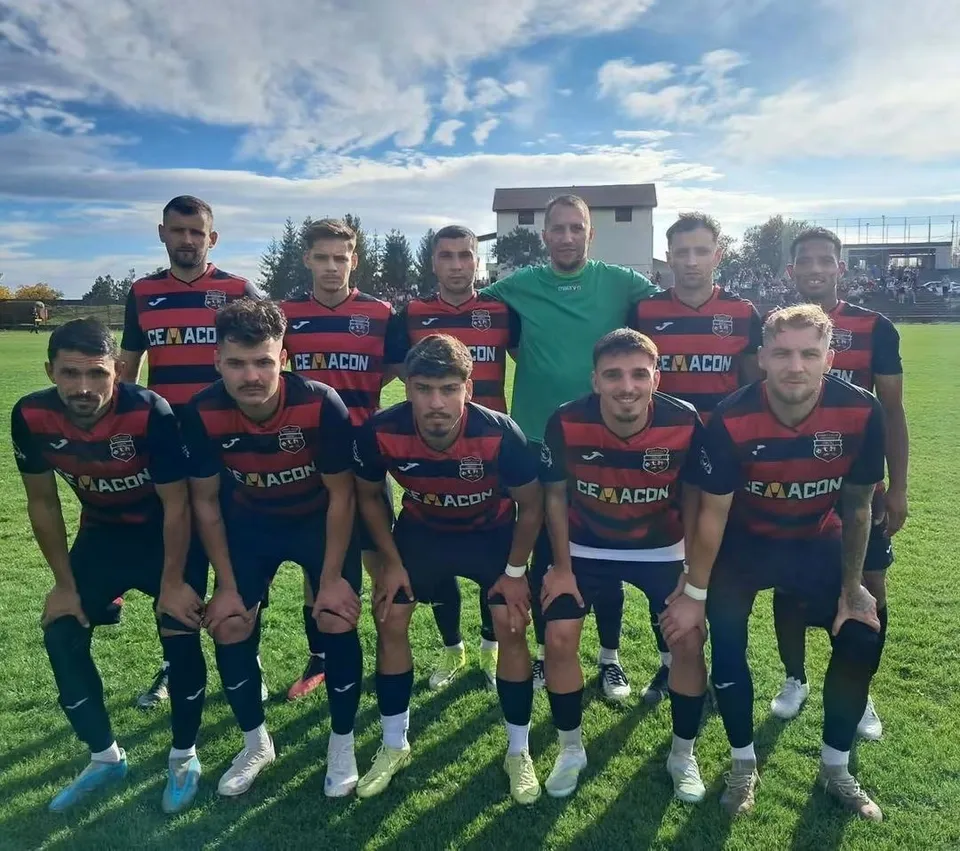 Un grup de fotbaliști în echipamente de sport roșii și negre pozează pe un teren de fotbal sub un cer cu nori.