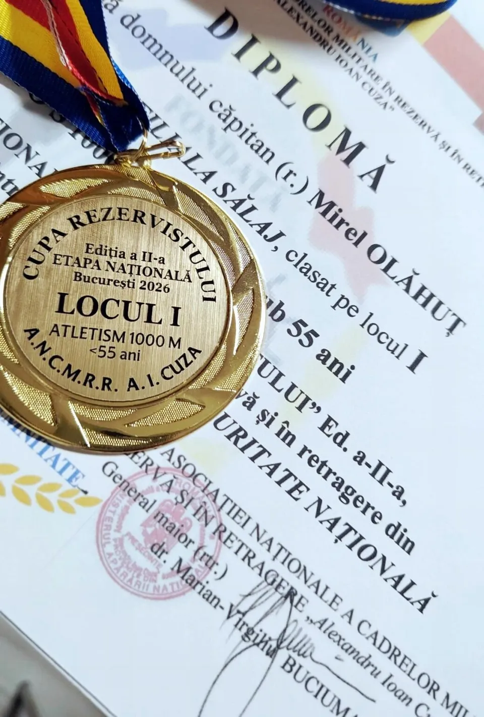 O medalie aurie cu inscripții este așezată pe o diplomă albă cu text negru și sigiliu roz.