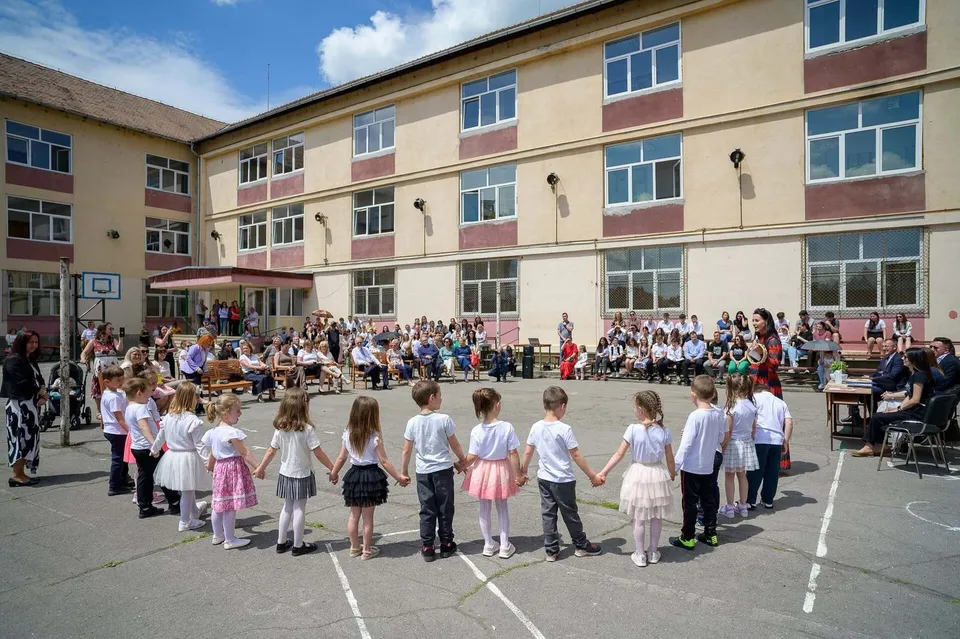 Un grup de copii stă în cerc pe o curte de școală în fața unei clădiri cu mai multe etaje, în timp ce adulți stau pe scaune în fundal.