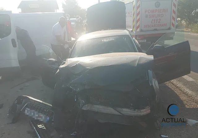 Un autoturism negru cu partea frontală zdrobită este situat pe drum, lângă un vehicul alb și o mașină neagră cu portiera deschisă.