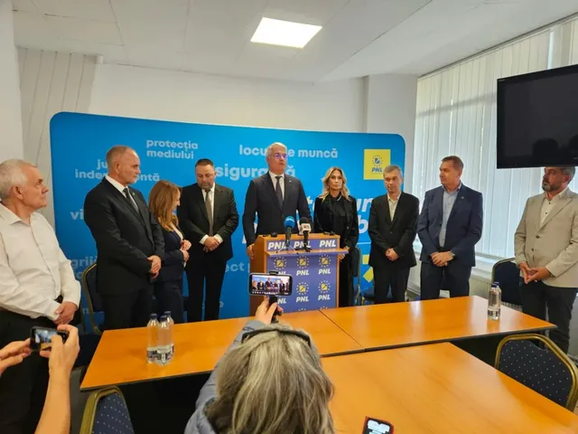 Un grup de oameni stă în picioare în fața unui panou albastru cu text și logo-uri, lângă o masă de lemn pe care sunt amplasate microfoane.