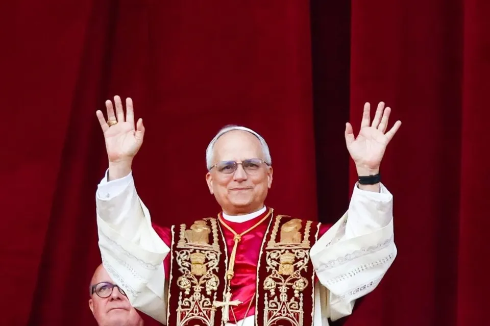 Un bătrân în haine liturgice albe și roșii cu broderii aurii ridică ambele mâini în sus în fața unui fundal de draperii roșii.