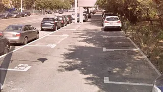 Mașini sunt parcate în locuri marcate cu linii albe pe o suprafață asfaltată lângă un drum cu trafic.