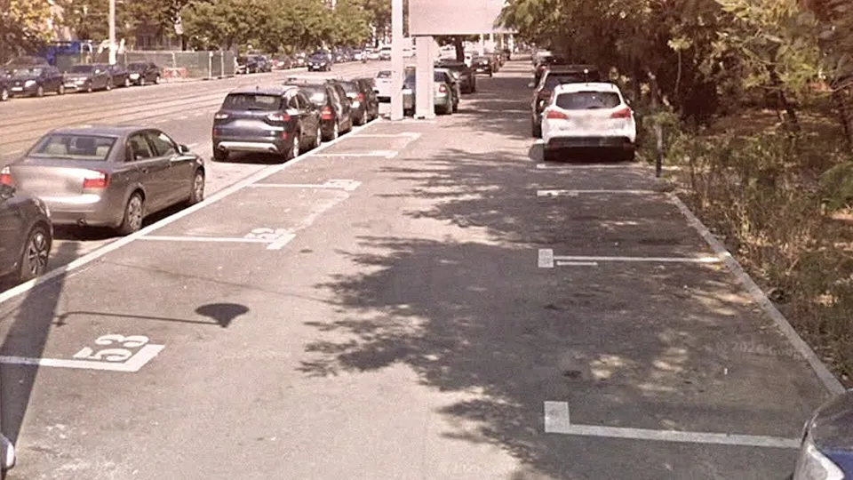Mașini sunt parcate în locuri marcate cu linii albe pe o suprafață asfaltată lângă un drum cu trafic.