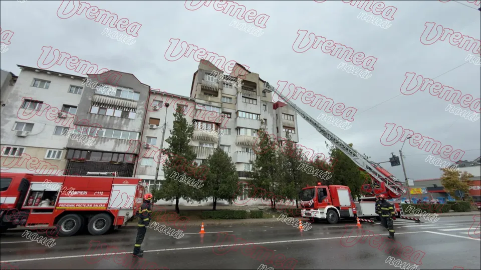 Pompierii lucrează lângă un camion roșu și o scară extensibilă în fața unui bloc de apartamente de culoare deschisă sub un cer gri.