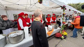 Oamenii în vestimentație roșie și albă servesc mâncare de la mese acoperite cu fețe de masă albe sub un cort alb.