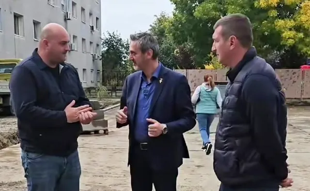 Trei bărbați stau în picioare pe un teren deschis lângă un clădire gri și un copac.