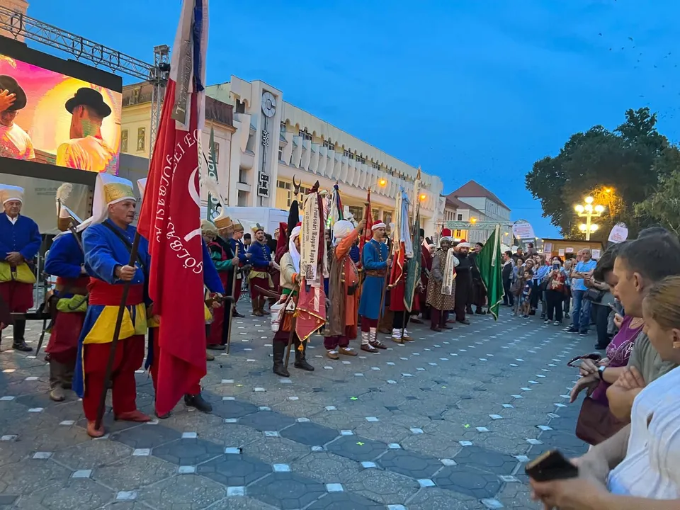 O procesiune de oameni îmbrăcați în costume colorate de epocă pășește pe o stradă pavată, purtând steaguri mari, în fața unui grup de spectatori.