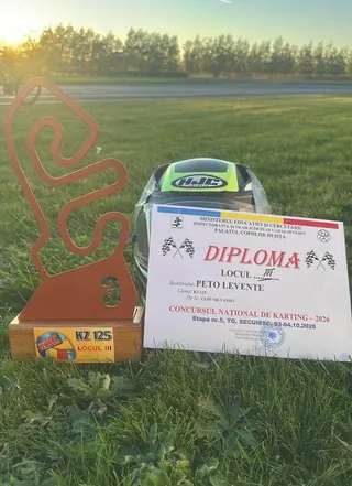 Un trofeu de lemn cu o formă sinuoasă, o diplomă albă și o cască de motociclist neagră cu detalii verzi sunt așezate pe iarbă.