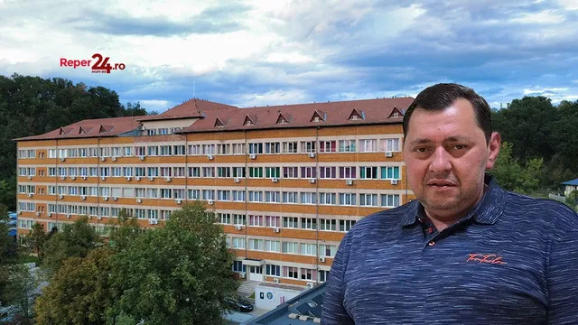 Un bărbat cu tricou polo albastru stă în prim-plan în fața unei clădiri mari cu fațadă portocalie și acoperiș roșu.