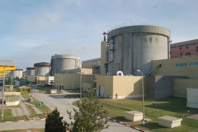 Două structuri cilindrice masive de beton gri sunt situate lângă clădiri industriale de culoare bej sub un cer senin.