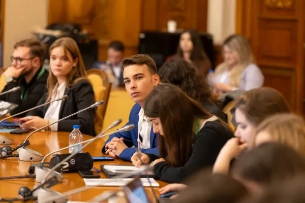 Un grup de tineri stă la o masă de conferință cu microfoane, cărți și sticle de apă într-o încăpere cu panouri de lemn în fundal.