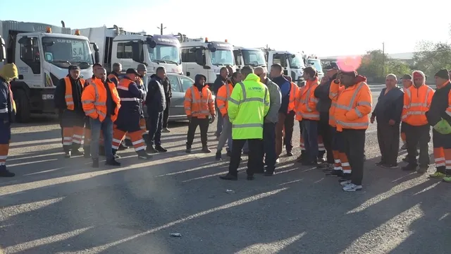 Un grup de persoane purtând veste reflectorizante portocalii și galbenii stă adunat pe o suprafață asfaltată lângă mai multe camioane albe.